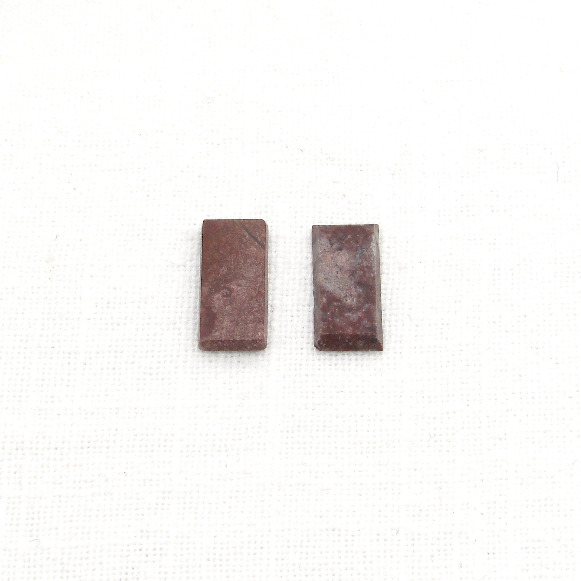 Dark red jasper cabochons are flat rectangles measuring approximately 16.3x8mm and 2.3.mm thick with beveled sides. Each opaque, polished stone is unique with inclusions forming patches and/or veins of beige, gray, and black.  Lighter-colored areas have more translucency than the rest of the stone.