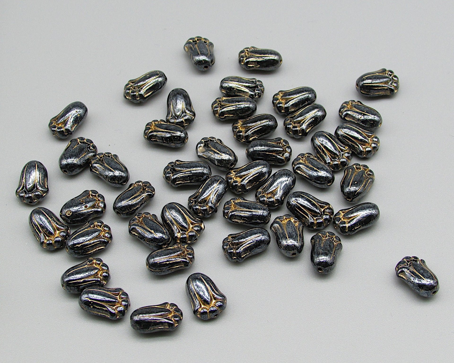 Opaque dark gray tulip-shaped pressed glass beads measure 12mm long, 8mm wide, 5.5mm thick. Center drilled lengthwise holes have an approximate diameter of 0.8mm. Beads are a shiny dark gray with a gold wash that highlights the details.