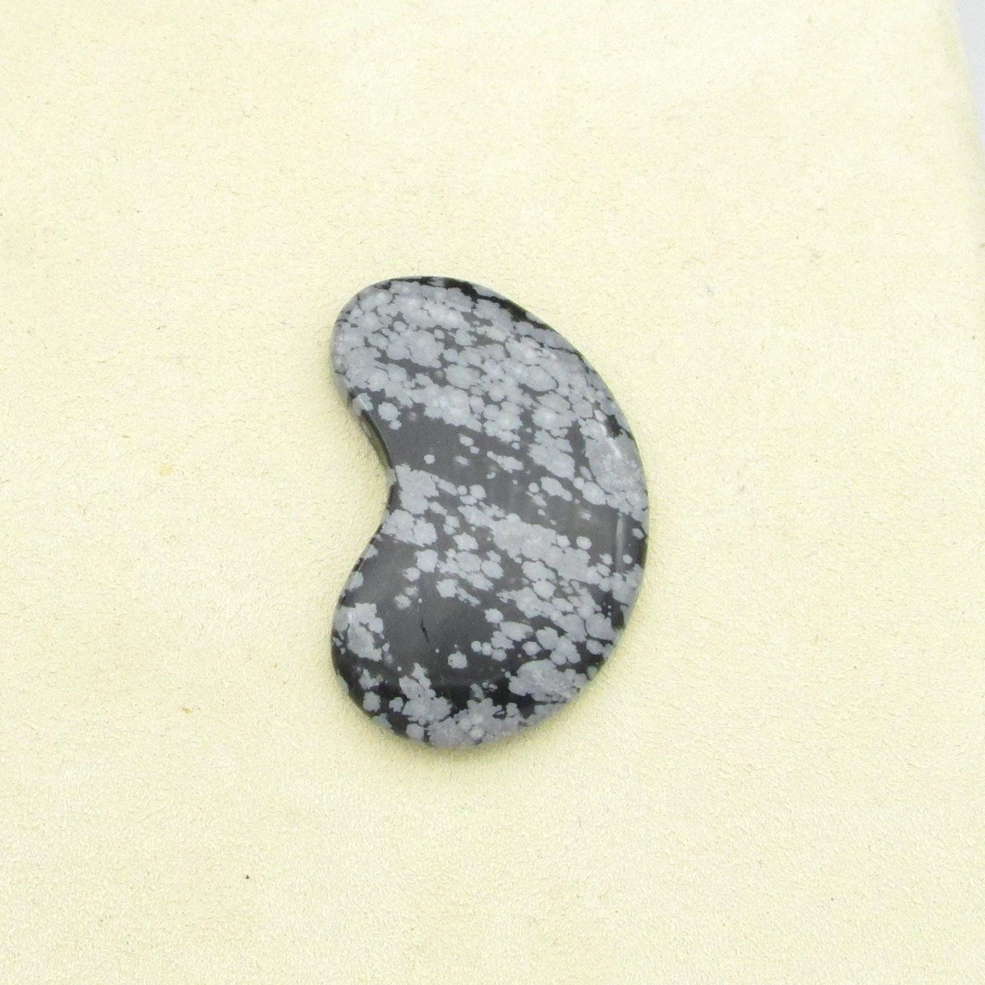Snowflake obsidian cabochon is kidney-shaped opaque black stone with gray blotches. Smoothly polished gemstone measures 40mm long, 22mm wide, and 4mm thick, with a weight of approximately 33.80 carats.