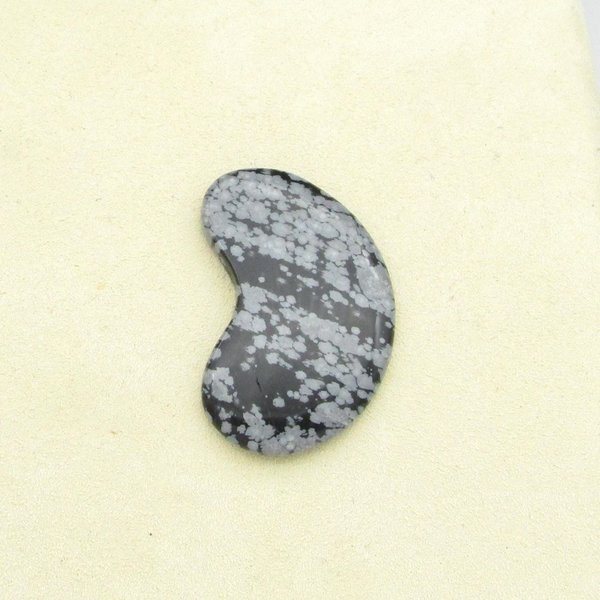 Snowflake obsidian cabochon is kidney-shaped opaque black stone with gray blotches. Smoothly polished gemstone measures 40mm long, 22mm wide, and 4mm thick, with a weight of approximately 33.80 carats.